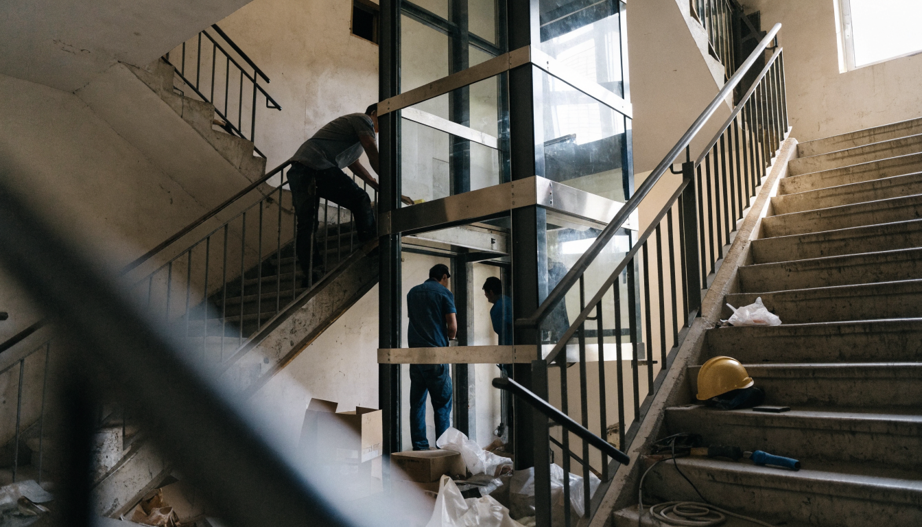 Instalación de un ascensor moderno en el hueco de la escalera de un edificio residencial, con operarios trabajando en la cabina metálica y luz natural entrando por un lucernario